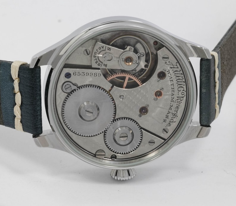 May include: A silver-toned wristwatch with a visible mechanical movement and a leather strap. The watch face displays intricate gears, springs, and the text "A.W.Co. Riverside, Waltham, Mass." The watch has a serial number 6559989.