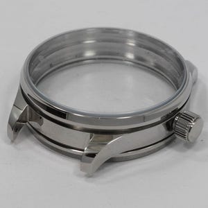 May include: A stainless steel watch case with a clear crystal. The case has a round shape with a crown on the side and lugs for attaching a watch band. The watch case is a component for watchmaking.