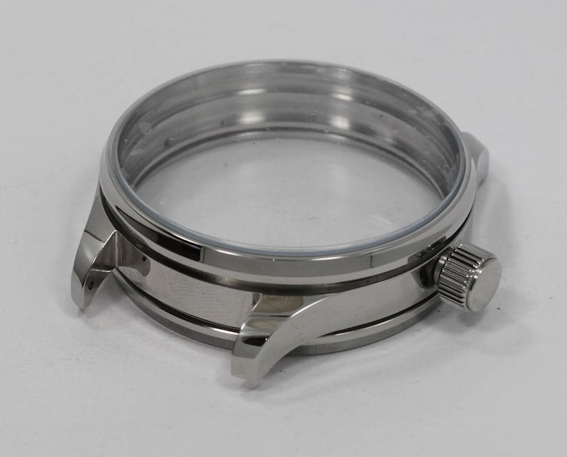 May include: A stainless steel watch case with a clear crystal. The case has a round shape with a crown on the side and lugs for attaching a watch band. The watch case is a component for watchmaking.