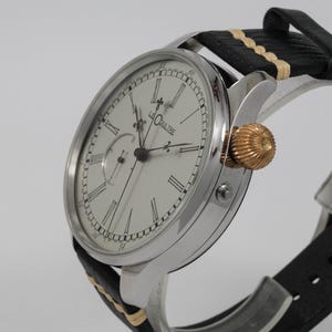May include: A silver wristwatch with a white face and black Roman numeral hour markers. The watch has a small subdial at the 3 o'clock position and the words "Le Coultre" printed on the face. The watch is on a black leather strap with a white stitching detail.