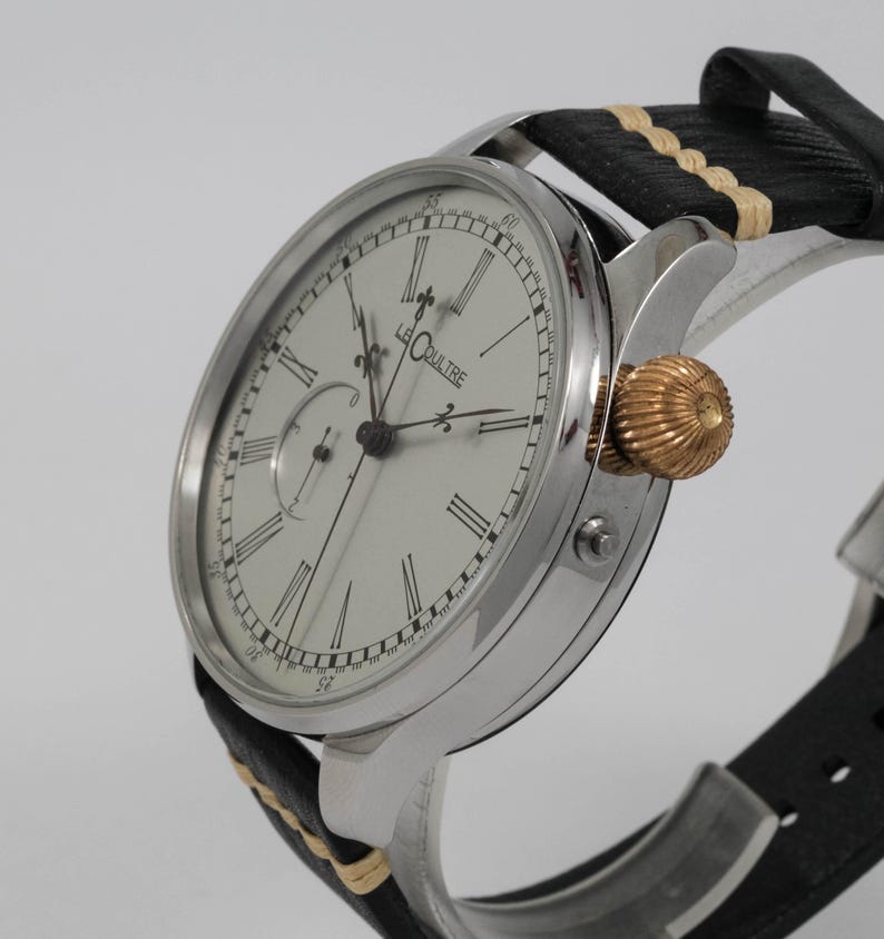 May include: A silver wristwatch with a white face and black Roman numeral hour markers. The watch has a small subdial at the 3 o'clock position and the words "Le Coultre" printed on the face. The watch is on a black leather strap with a white stitching detail.