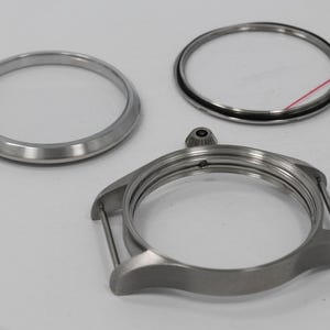 May include: A collection of watch components, including a silver-colored watch case, a bezel, and a crystal ring. The watch case has lugs for attaching a strap. The parts are arranged on a white surface.