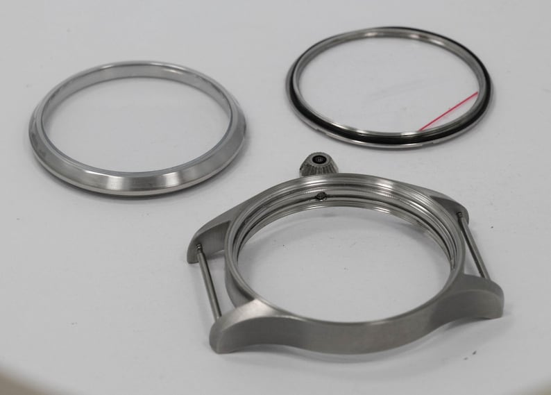 May include: A collection of watch components, including a silver-colored watch case, a bezel, and a crystal ring. The watch case has lugs for attaching a strap. The parts are arranged on a white surface.