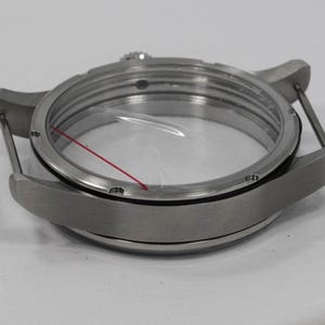 May include: A stainless steel watch case with a clear crystal and a red line. The watch case has a round shape with lugs for attaching a strap. The watch case is designed to protect the watch movement.