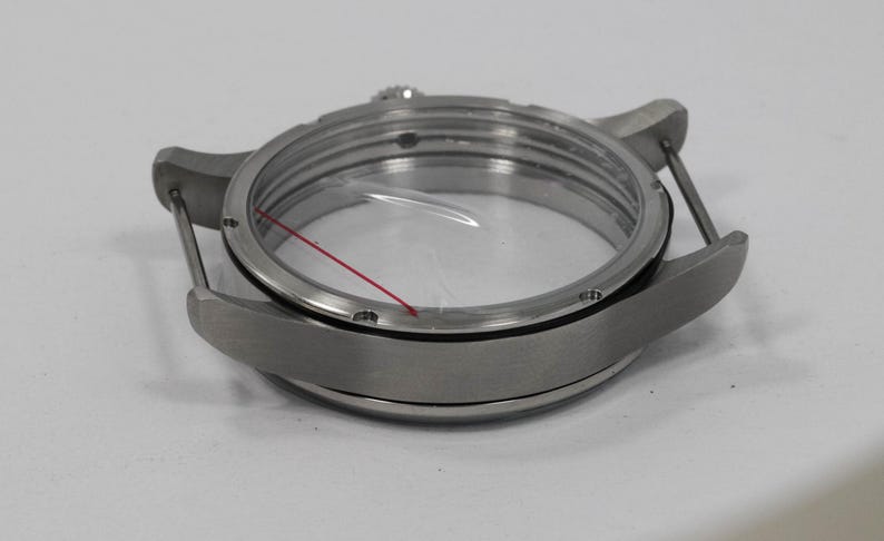 May include: A stainless steel watch case with a clear crystal and a red line. The watch case has a round shape with lugs for attaching a strap. The watch case is designed to protect the watch movement.