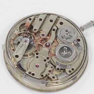 Puede incluir: Primer plano de un movimiento de reloj antiguo, que muestra intrincados engranajes, palancas y joyas. Los componentes plateados est&aacute;n dispuestos en un marco circular, con una tija de cuerda que se extiende lateralmente. El dise&ntilde;o resalta la precisi&oacute;n y la artesan&iacute;a de los relojes antiguos.