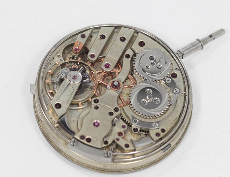 Puede incluir: Primer plano de un movimiento de reloj antiguo, que muestra intrincados engranajes, palancas y joyas. Los componentes plateados est&aacute;n dispuestos en un marco circular, con una tija de cuerda que se extiende lateralmente. El dise&ntilde;o resalta la precisi&oacute;n y la artesan&iacute;a de los relojes antiguos.