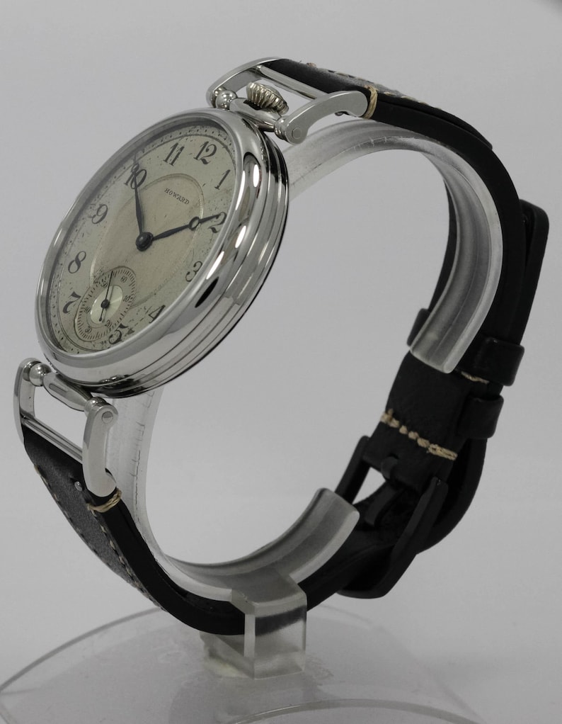 K&ouml;nnte beinhalten: Eine silberfarbene Armbanduhr mit rundem Geh&auml;use und cremefarbenem Zifferblatt mit schwarzen arabischen Ziffern und dem Schriftzug Howard. Die Uhr hat d&uuml;nne schwarze Zeiger, ein kleines Hilfszifferblatt auf der Sechs-Uhr-Position und ein schwarzes Lederarmband.