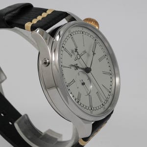 May include: A silver wristwatch with a white face and black hands. The watch has Roman numeral hour markers and a small seconds dial at the 6 o'clock position. The watch is on a black leather strap with a cream-colored stitching.