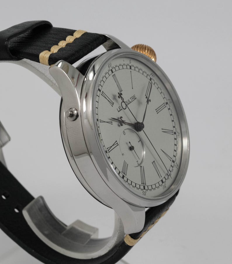 May include: A silver wristwatch with a white face and black hands. The watch has Roman numeral hour markers and a small seconds dial at the 6 o'clock position. The watch is on a black leather strap with a cream-colored stitching.