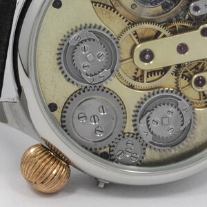 May include: A close-up of a silver pocket watch with a clear face revealing the intricate inner workings of the watch mechanism. The watch has a gold-toned crown and a black leather strap.