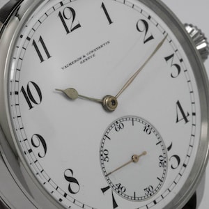 Puede incluir: Un reloj de pulsera anal&oacute;gico blanco con marcadores de hora en n&uacute;meros romanos negros y marcadores de minutos negros. La esfera del reloj tiene un peque&ntilde;o subdial en la posici&oacute;n de las 6 en punto. La esfera del reloj tambi&eacute;n tiene el texto "VACHERON & CONSTANTIN GENEVE".