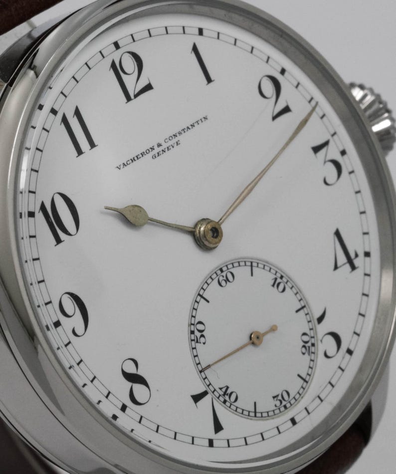 Puede incluir: Un reloj de pulsera anal&oacute;gico blanco con marcadores de hora en n&uacute;meros romanos negros y marcadores de minutos negros. La esfera del reloj tiene un peque&ntilde;o subdial en la posici&oacute;n de las 6 en punto. La esfera del reloj tambi&eacute;n tiene el texto "VACHERON & CONSTANTIN GENEVE".