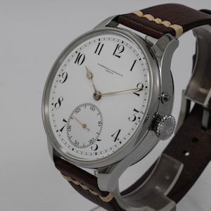 Puede incluir: Un reloj de pulsera plateado con una esfera blanca y marcadores de hora romanos negros. El reloj tiene un peque&ntilde;o subdial para los segundos y una correa de cuero marr&oacute;n con costuras blancas.