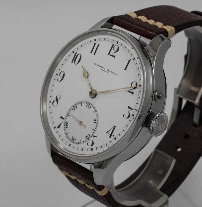 Puede incluir: Un reloj de pulsera plateado con una esfera blanca y marcadores de hora romanos negros. El reloj tiene un peque&ntilde;o subdial para los segundos y una correa de cuero marr&oacute;n con costuras blancas.