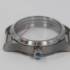 May include: A stainless steel watch case with a clear crystal and a blue-accented crown. The watch case has a round face and is designed to hold the watch movement and dial. The lugs are curved for a comfortable fit.