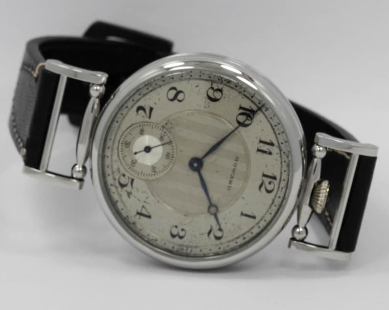 K&ouml;nnte beinhalten: Eine silberfarbene Armbanduhr mit rundem Geh&auml;use und schwarzem Lederarmband. Das cremefarbene Zifferblatt zeigt schwarze arabische Ziffern, blaue Zeiger, ein kleines Sekunden-Hilfszifferblatt und den Schriftzug HOWARD in Schwarz auf der rechten Seite.