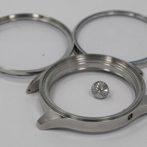 May include: A disassembled watch case with three silver components. The watch case, two bezels, and a crown are displayed on a white surface. The watch case has two lugs and a small hole for the crown.