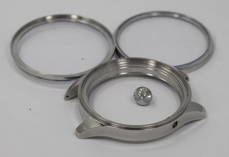 May include: A disassembled watch case with three silver components. The watch case, two bezels, and a crown are displayed on a white surface. The watch case has two lugs and a small hole for the crown.