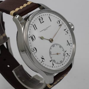 Puede incluir: Un reloj de pulsera Vacheron Constantin plateado con una esfera blanca y marcadores de hora romanos negros. El reloj tiene un peque&ntilde;o subdial para los segundos y una correa de cuero marr&oacute;n.