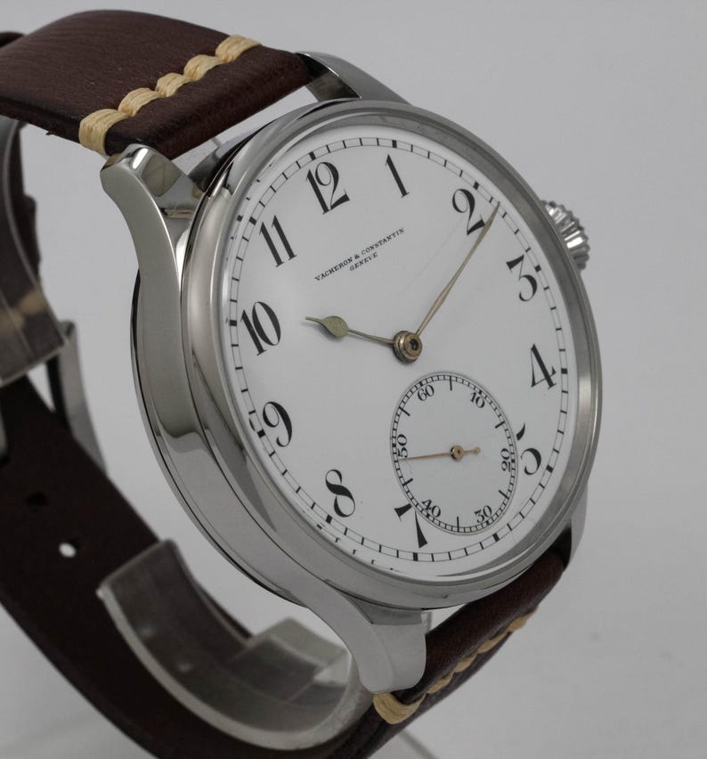 Puede incluir: Un reloj de pulsera Vacheron Constantin plateado con una esfera blanca y marcadores de hora romanos negros. El reloj tiene un peque&ntilde;o subdial para los segundos y una correa de cuero marr&oacute;n.