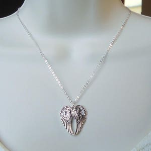 K&ouml;nnte beinhalten: Eine silberne Kettenkette mit einem Anh&auml;nger in Form von zwei Engelsfl&uuml;geln. Die Fl&uuml;gel sind mit komplizierten Designs verziert und in der Mitte verbunden.