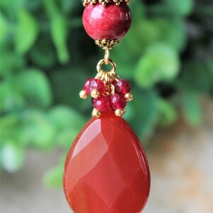 May include: A pendant featuring a large, faceted, teardrop-shaped red stone suspended from a cluster of smaller, round, deep red beads. The assembly is topped with a single, larger, smooth red bead encased in ornate, antique-gold metal filigree caps.