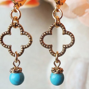 May include: A pair of gold-toned dangle earrings featuring a quatrefoil-shaped charm with a beaded texture. Each earring is suspended from a lever-back hook and finished with a small, round, turquoise-colored bead attached at the bottom.