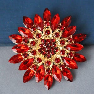 May include: A large red and gold brooch in the shape of a flower. The brooch is made of metal and rhinestones. The center of the brooch is a cluster of red rhinestones surrounded by a gold metal design. The outer petals of the brooch are made of red rhinestones.