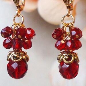May include: A pair of dangle earrings featuring clusters of small, faceted, deep red beads suspended from gold-toned metal hooks. Each earring includes a larger, faceted red bead at the base, topped with an ornate, gold-toned metal cap.
