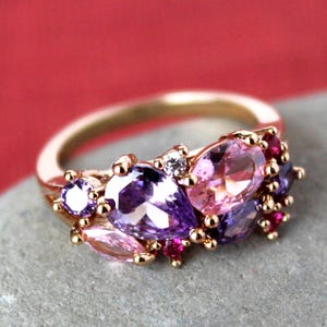 May include: A gold-toned ring featuring an array of colorful gemstones. The ring showcases a cluster of purple, pink, and red stones, with a small white stone accent. The gemstones are set on a gold band, creating a vibrant and eye-catching design.