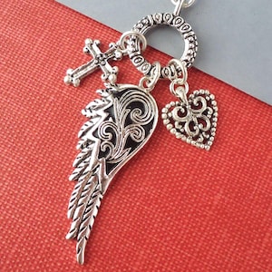 May include: Silver-toned pendant necklace featuring a cross, a wing with black inlay, and a heart charm. The charms are suspended from a decorative ring clasp. The necklace is displayed against a red and gray background.