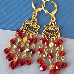 May include: A pair of gold-colored dangle earrings with red and gold beads. The earrings have a decorative top with a series of beaded strands hanging down. The red beads are faceted and the gold beads are a mix of shapes.