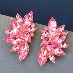 May include: A pair of pink crystal earrings with a floral design. Each earring features multiple marquise-cut crystals in varying shades of pink, arranged around a central cluster. The crystals are set in a gold-tone metal.