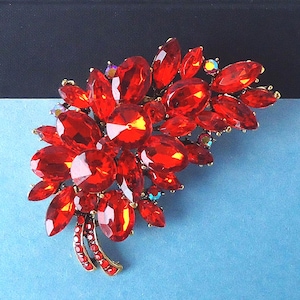 May include: A red crystal brooch in a floral design. The brooch features numerous faceted red crystals in various shapes, clustered together to resemble a bouquet. The setting is gold-toned, with additional smaller crystals.