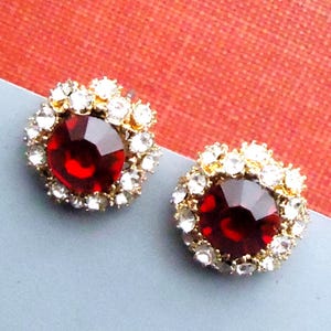 Op de afbeelding: Een paar oorstekers met grote, facetgeslepen, dieprode edelstenen, omringd door kleinere, heldere kristallen. De goudkleurige zettingen worden gepresenteerd op een lichtblauwe ondergrond tegen een rode achtergrond.