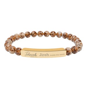 Op de afbeelding: Een armband met bruine en beige gespikkelde kralen. Een goudkleurige rechthoekige balk in het midden is gegraveerd met de woorden "Jehovah Jireh meets my needs". De armband staat op een witte achtergrond.