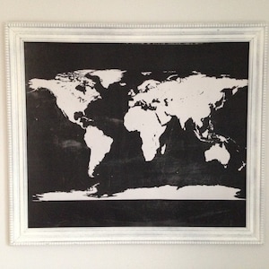 May include: A framed world map print. The map features a black background with white continents and is encased in a white, ornate frame. The artwork is a decorative piece suitable for home or office decor.
