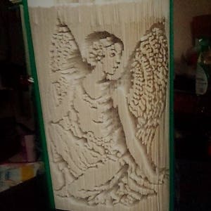 Può includere: Un libro bianco con le pagine piegate in una scultura 3D di un angelo con le ali. La scultura è in piedi su una superficie scura. Il testo "Creased Chronicles" è visibile nella parte inferiore dell'immagine.