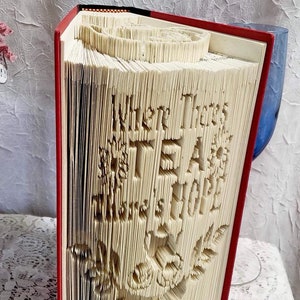 May include: A red book with the words "Creased Chronicles" on the spine. The pages of the book have been folded into a 3D design that creates the words "When There's Tea, There's Hope".