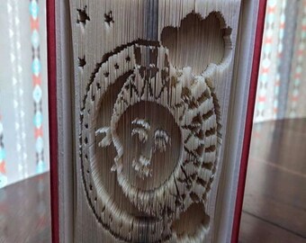 Può includere: Un libro con una copertina rossa è stato piegato in una scultura 3D di una luna crescente e un sole con un viso. La scultura è intitolata "Creased Chronicles".