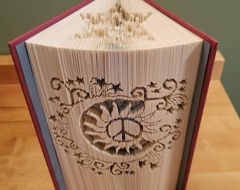 Può includere: Un libro con una copertina rossa è stato piegato in un design tridimensionale. Il design è una stella con un segno di pace al suo interno. Il libro è in piedi su una superficie di legno. Il testo "Creating Creases" è visibile nella parte inferiore dell'immagine.