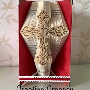 May include: A book folded into the shape of a cross with intricate details. The cross is made of white paper and is standing on a red base with white lace trim. The base has the text "Creating Creases" printed on it.