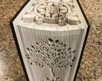 May include: A book with a white cover has been folded into a three-dimensional tree shape. The tree is detailed with intricate swirls and lines. The book is standing upright on a brown surface. The text "Creased Chronicles" is visible at the bottom of the image.