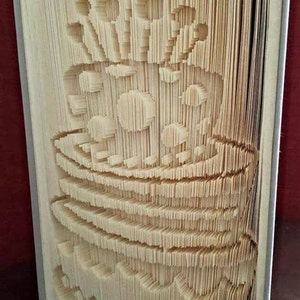 May include: A book art sculpture of a birthday cake with candles, made by folding the pages of a book. The sculpture is titled "Creased Chronicles".