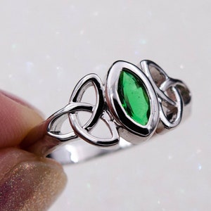 Pode incluir: Um anel de prata com uma pedra verde oval. O anel apresenta um design de nó celta em ambos os lados da pedra. O anel é segurado por um dedo.