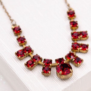 Op de afbeelding: Een vintage-stijl ketting met een reeks levendige rode edelstenen gezet in een goudkleurig metaal. De ketting heeft een delicate ketting en een symmetrisch ontwerp, met edelstenen van verschillende grootte. De ketting wordt gepresenteerd op een witte ondergrond.