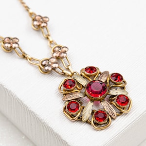 Könnte beinhalten: Eine Vintage-Halskette in Goldoptik mit einem floralen Anhänger. Der Anhänger hat einen großen, runden, roten Edelstein, der von kleineren roten Edelsteinen und goldfarbenen Blättern umgeben ist. Die Halskette hat eine zarte Kette und kleine Blumenakzente.