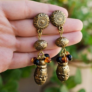 May include: A pair of antique gold earrings. Each earring has a circular top with a sunburst design, connected by chains to an oval bead, a small cluster of beads, and a decorative, urn-shaped bottom.