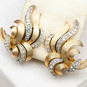 Può includere: Orecchini a clip color oro con un design a spirale e strutturato. Gli orecchini presentano una serie di elementi curvi, accentuati da strass chiari e scintillanti. Gli orecchini a clip sono esposti su una superficie bianca.